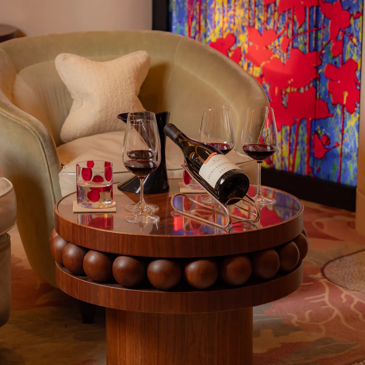 Wine bottle and two glasses on a round table beside an armchair and colourful artwork.