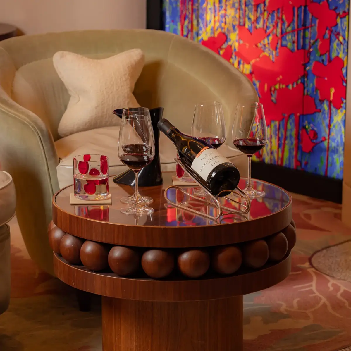 Wine bottle and two glasses on a round table beside an armchair and colourful artwork.