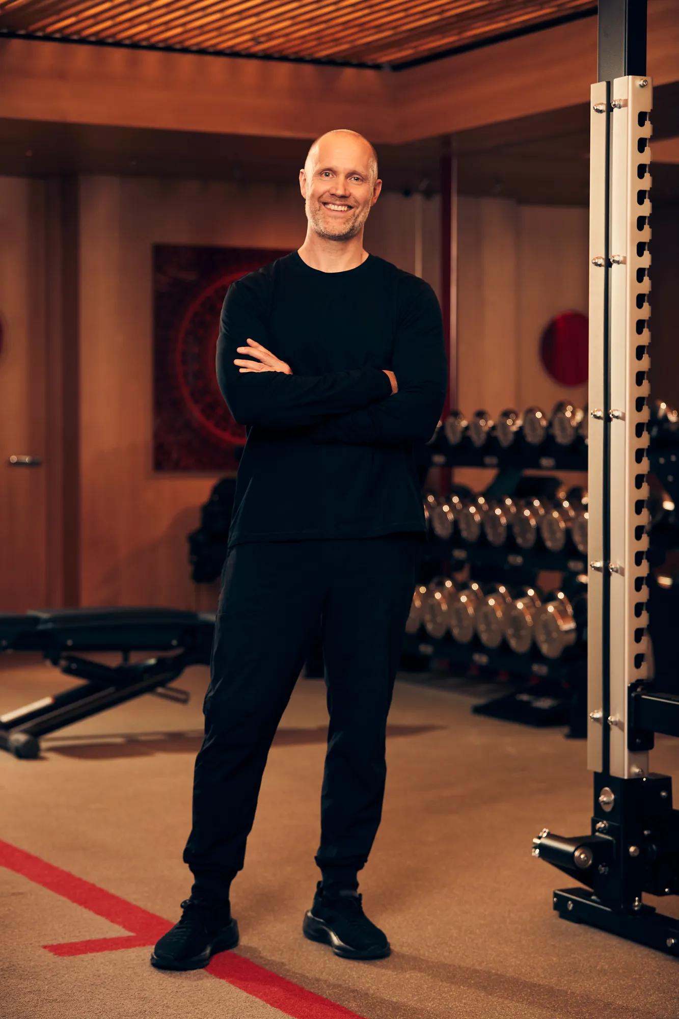 Smiling man in black sportswear stands with arms crossed in a modern gym with weights and equipment behind.