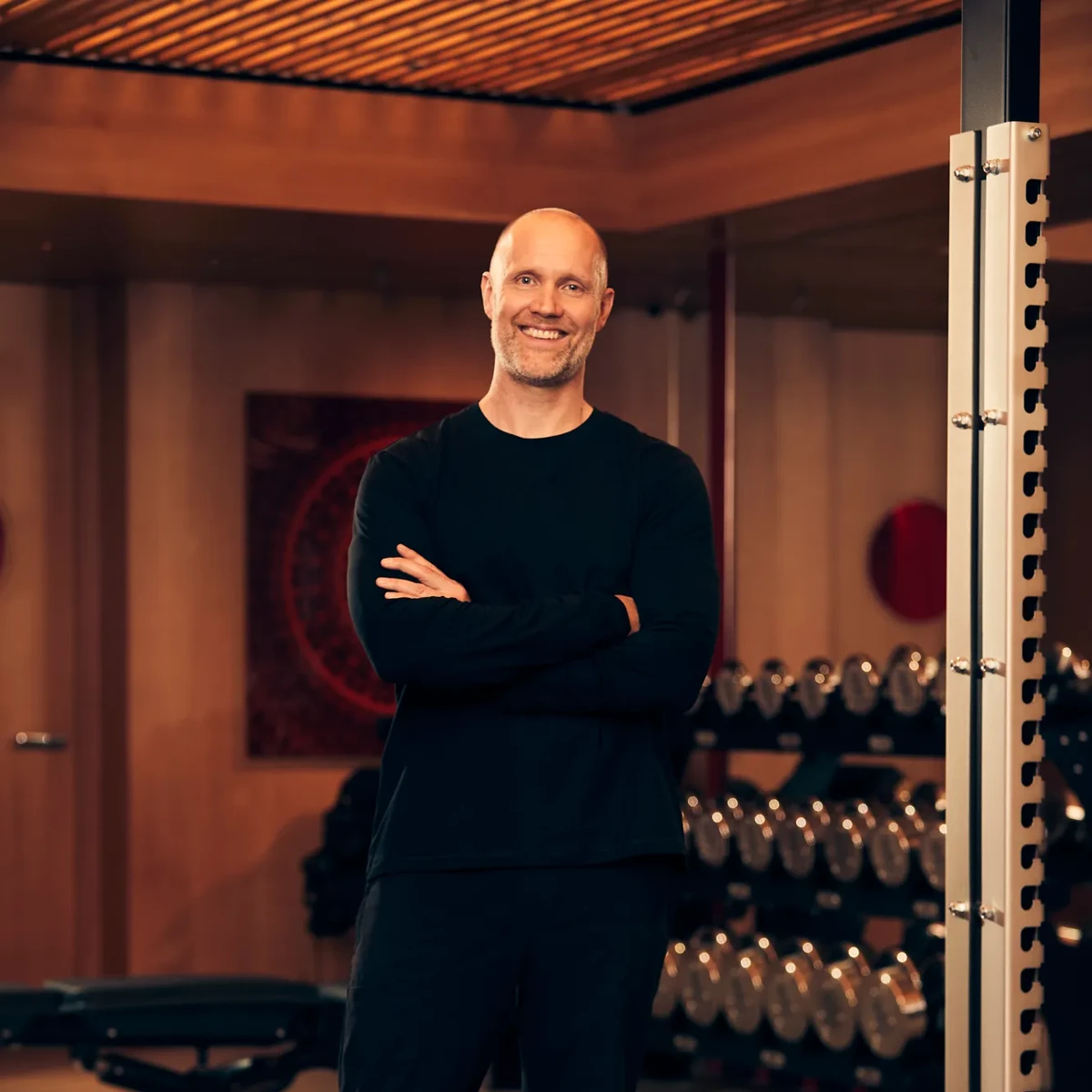 Smiling man in black sportswear stands with arms crossed in a modern gym with weights and equipment behind.