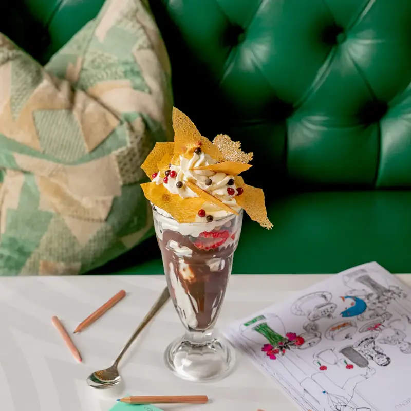 Chocolate sundae served in a tall glass, topped with whipped cream, honeycomb pieces, and red berries. The dessert sits on a white table beside coloring pencils, a children’s menu, and a mint-green Claridge’s chocolate bar, against a green tufted banquette.