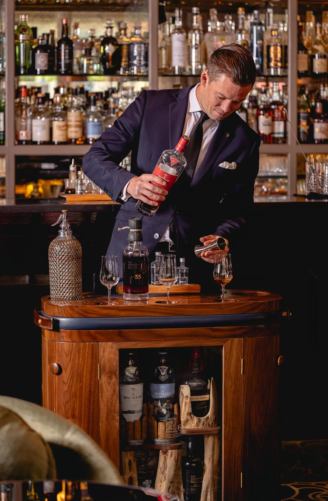 Smartly dressed bartender pours a spirit from a decanter at a wooden drinks trolley in front of a wall of bottles.