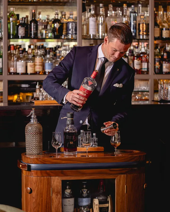 Smartly dressed bartender pours a spirit from a decanter at a wooden drinks trolley in front of a wall of bottles.