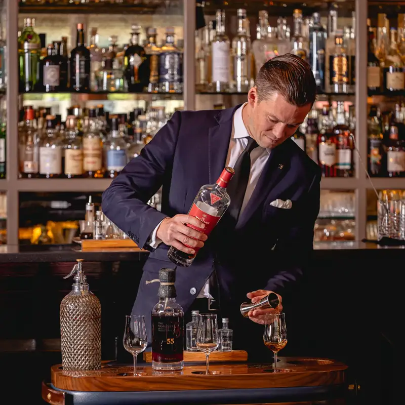Smartly dressed bartender pours a spirit from a decanter at a wooden drinks trolley in front of a wall of bottles.