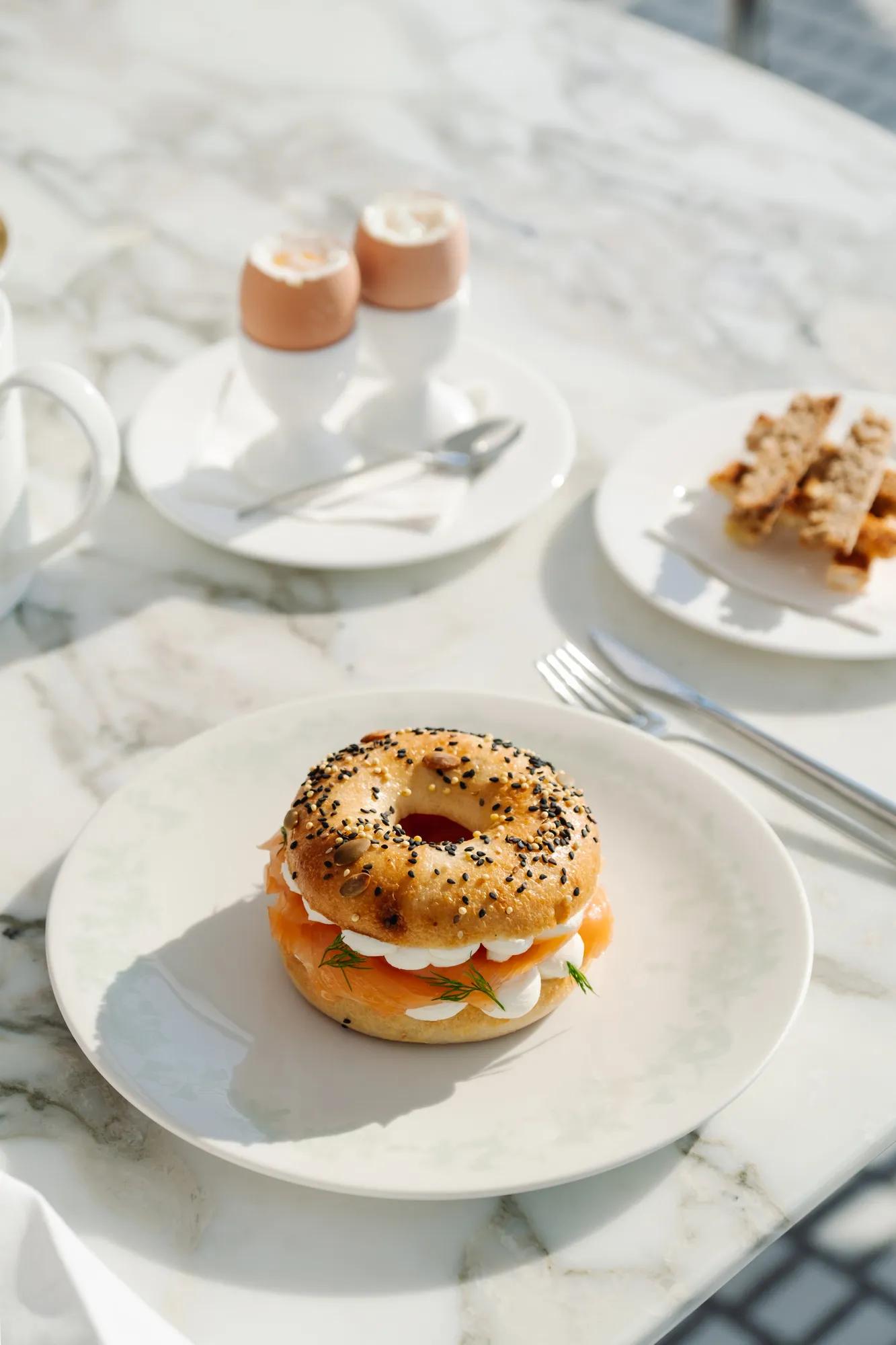 Breakfast table with a sesame and poppy seed bagel filled with smoked salmon, cream cheese, and herbs, served alongside soft-boiled eggs and toast soldiers.
