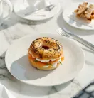 Breakfast table with a sesame and poppy seed bagel filled with smoked salmon, cream cheese, and herbs, served alongside soft-boiled eggs and toast soldiers.
