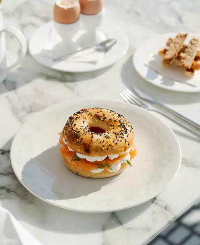 Table de petit-déjeuner avec un bagel aux graines de sésame et de pavot garni de saumon fumé, fromage frais et herbes, accompagné d’œufs à la coque et de mouillettes.