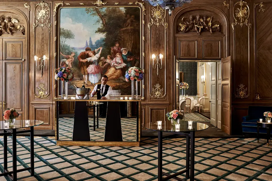 A bartender stands behind a mirrored bar in Claridge’s French Salon, surrounded by dark wood panelling, gilded details, floral displays, and an ornate mural depicting a pastoral scene.