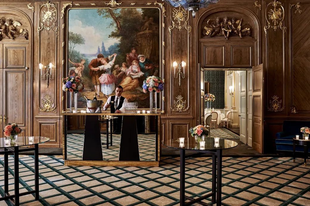 A bartender stands behind a mirrored bar in Claridge’s French Salon, surrounded by dark wood panelling, gilded details, floral displays, and an ornate mural depicting a pastoral scene.