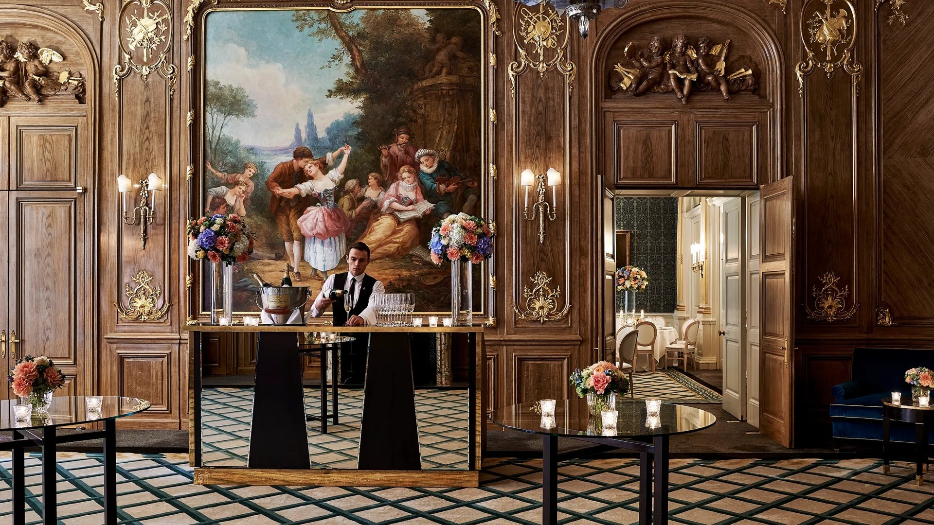 A bartender stands behind a mirrored bar in Claridge’s French Salon, surrounded by dark wood panelling, gilded details, floral displays, and an ornate mural depicting a pastoral scene.