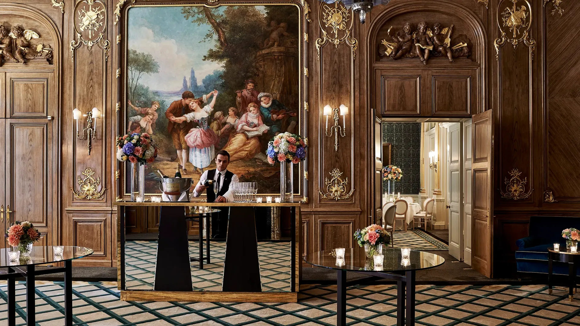 A bartender stands behind a mirrored bar in Claridge’s French Salon, surrounded by dark wood panelling, gilded details, floral displays, and an ornate mural depicting a pastoral scene.