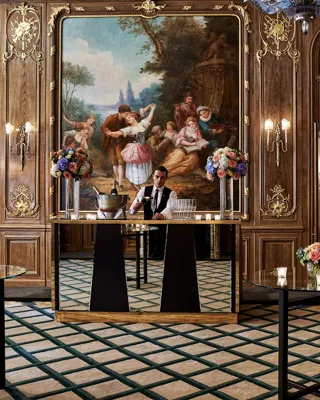 A bartender stands behind a mirrored bar in Claridge’s French Salon, surrounded by dark wood panelling, gilded details, floral displays, and an ornate mural depicting a pastoral scene.