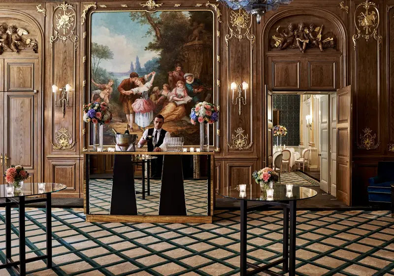 A bartender stands behind a mirrored bar in Claridge’s French Salon, surrounded by dark wood panelling, gilded details, floral displays, and an ornate mural depicting a pastoral scene.