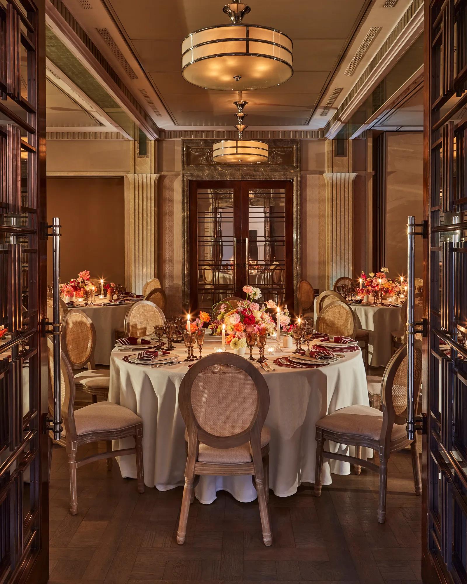 Warmly lit dining room with round tables set with floral centerpieces and candles, surrounded by elegant wooden chairs.