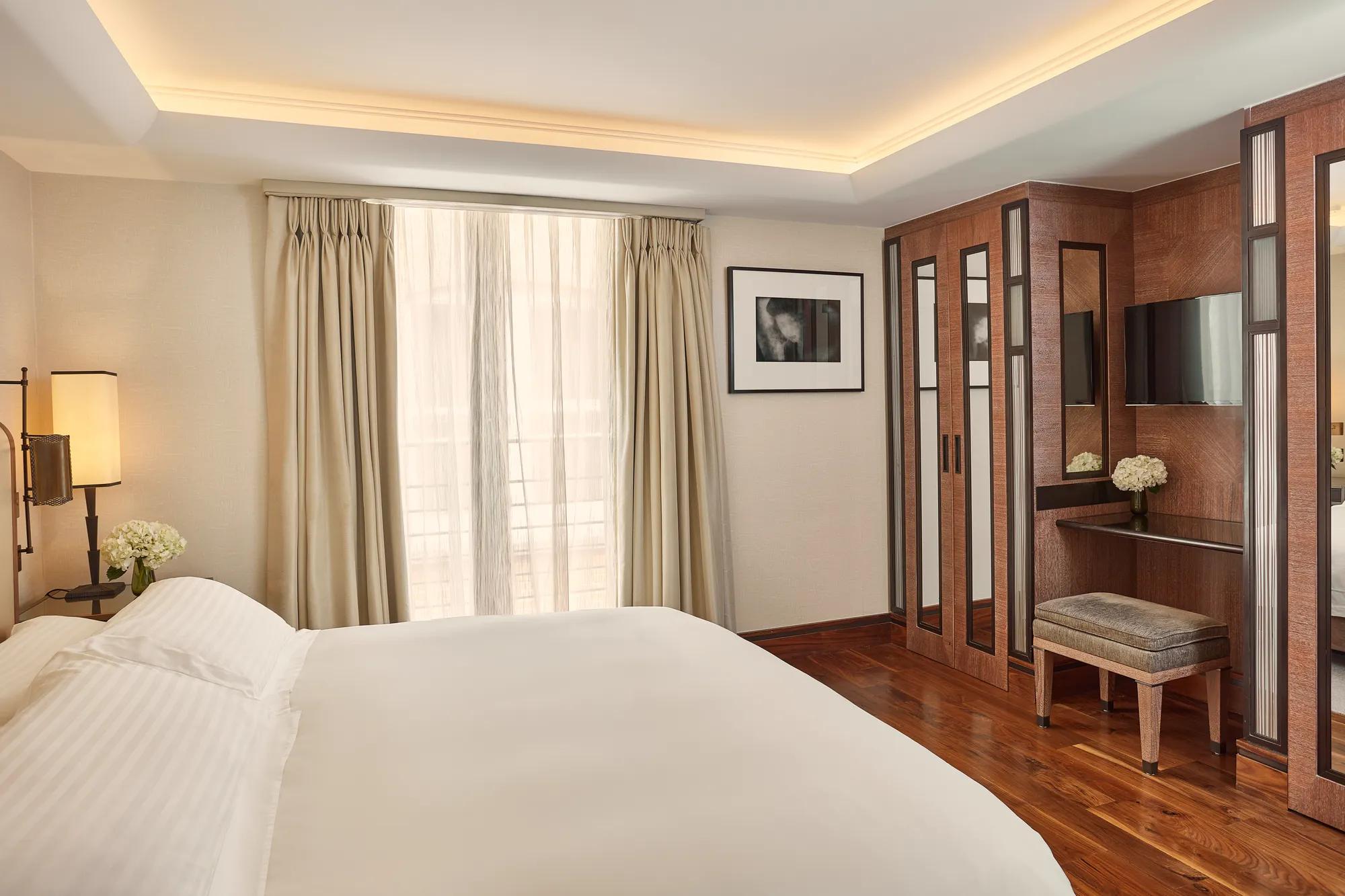 Bright bedroom with a neatly made bed, floor-to-ceiling curtains, wooden wardrobes, and a small vanity with a stool and flowers.