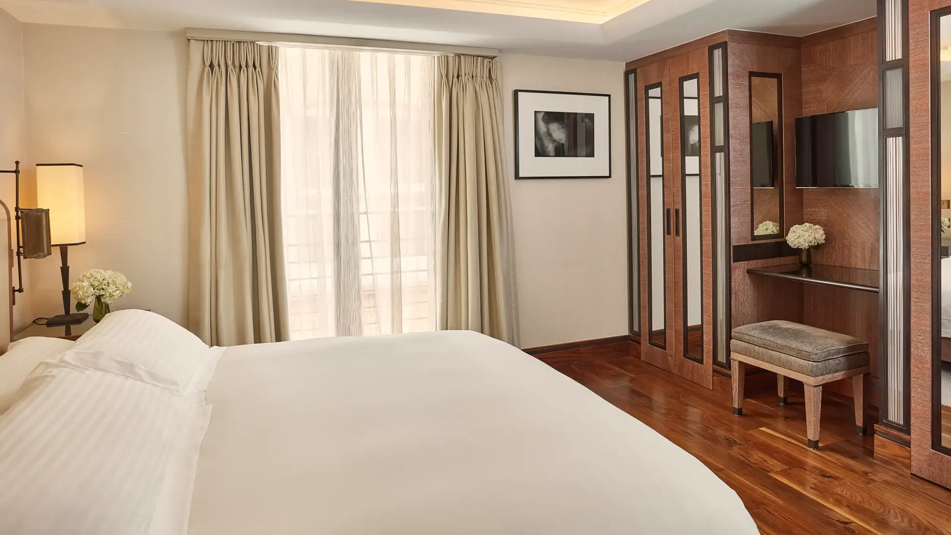 Bright bedroom with a neatly made bed, floor-to-ceiling curtains, wooden wardrobes, and a small vanity with a stool and flowers.