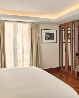 Bright bedroom with a neatly made bed, floor-to-ceiling curtains, wooden wardrobes, and a small vanity with a stool and flowers.