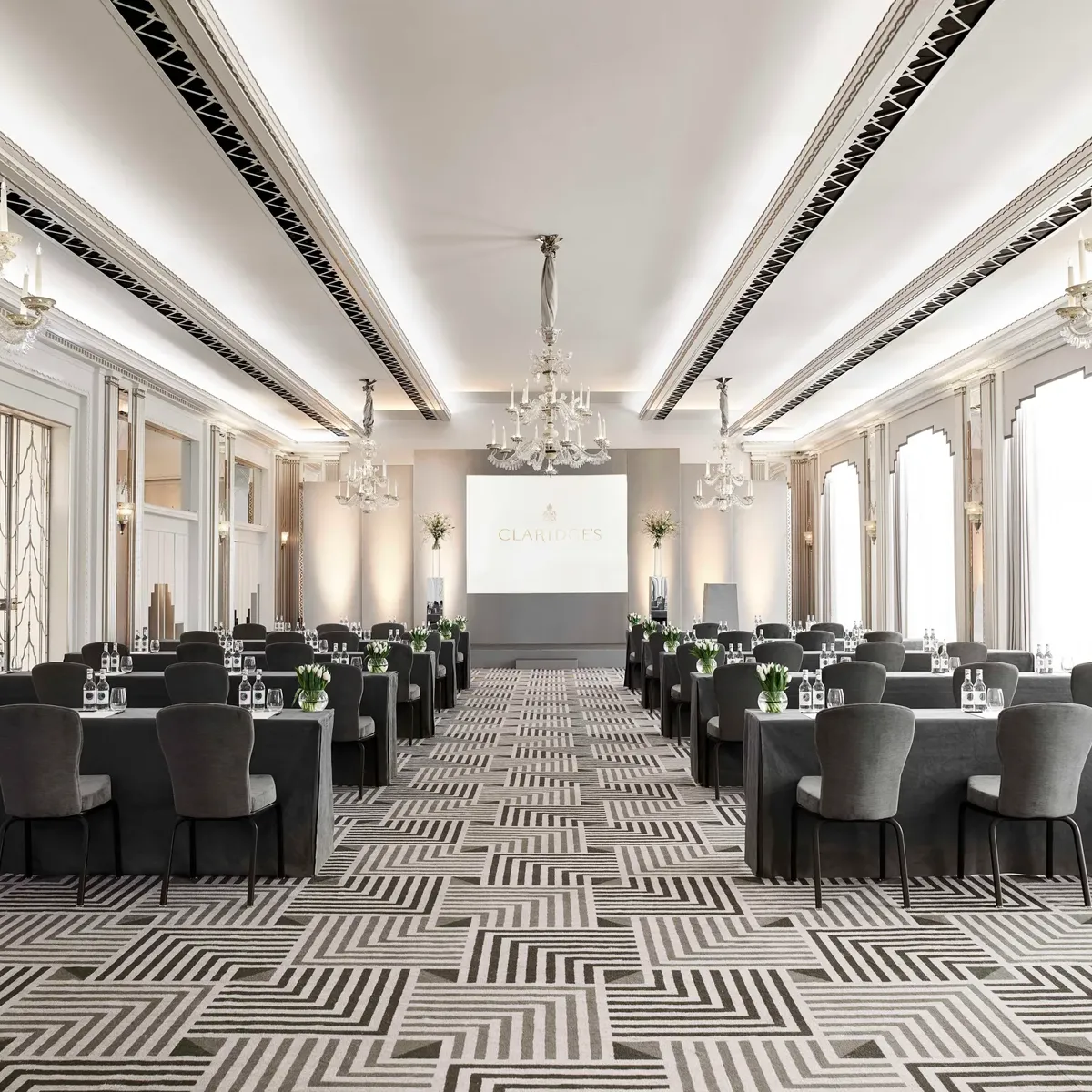 The Ballroom at Claridge’s arranged in a conference setup with long grey-covered tables and chairs facing a presentation screen, lit by chandeliers and large arched windows.