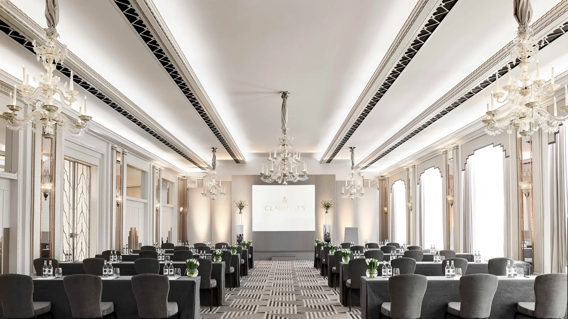 The Ballroom at Claridge’s arranged in a conference setup with long grey-covered tables and chairs facing a presentation screen, lit by chandeliers and large arched windows.