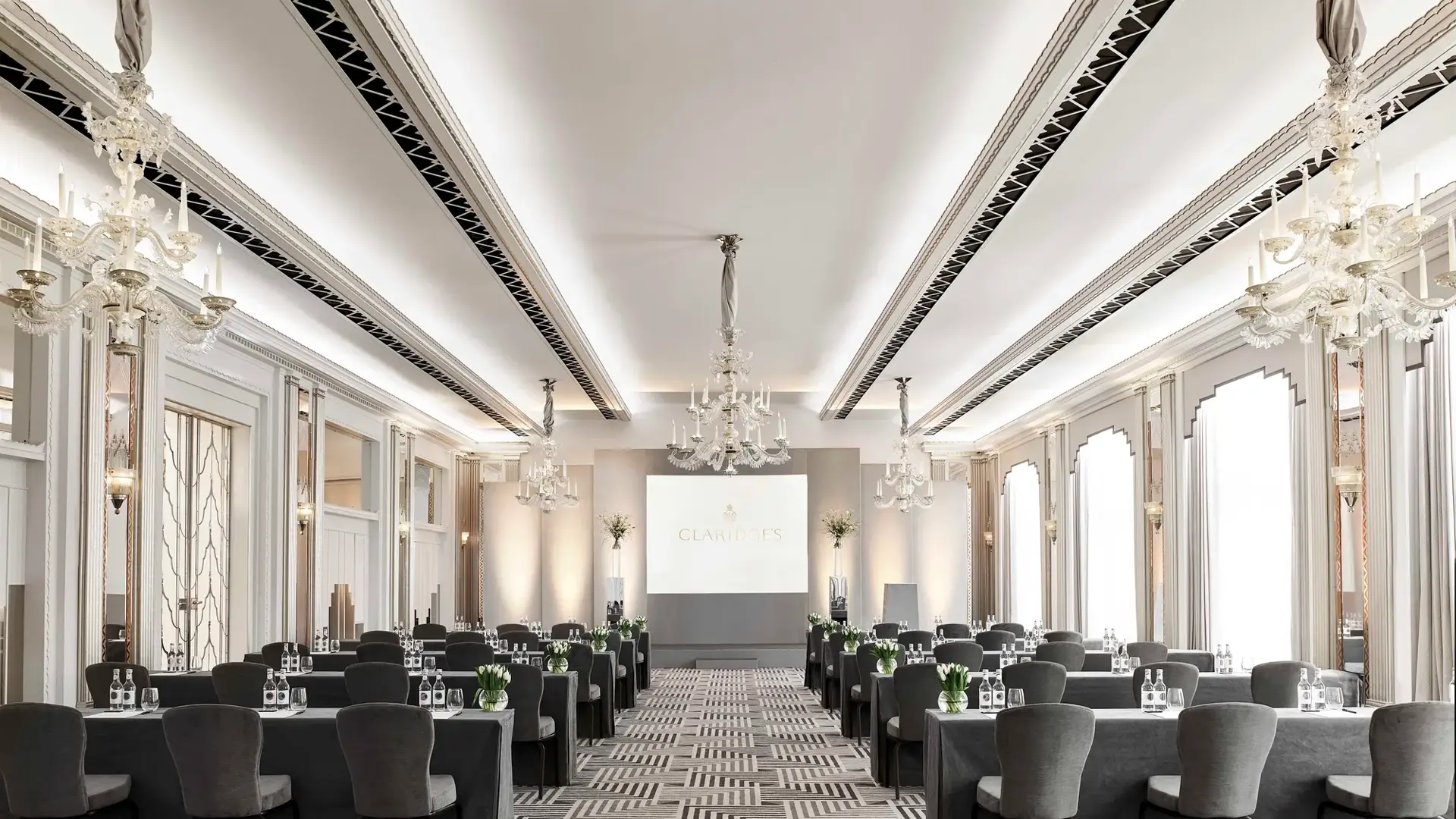The Ballroom at Claridge’s arranged in a conference setup with long grey-covered tables and chairs facing a presentation screen, lit by chandeliers and large arched windows.