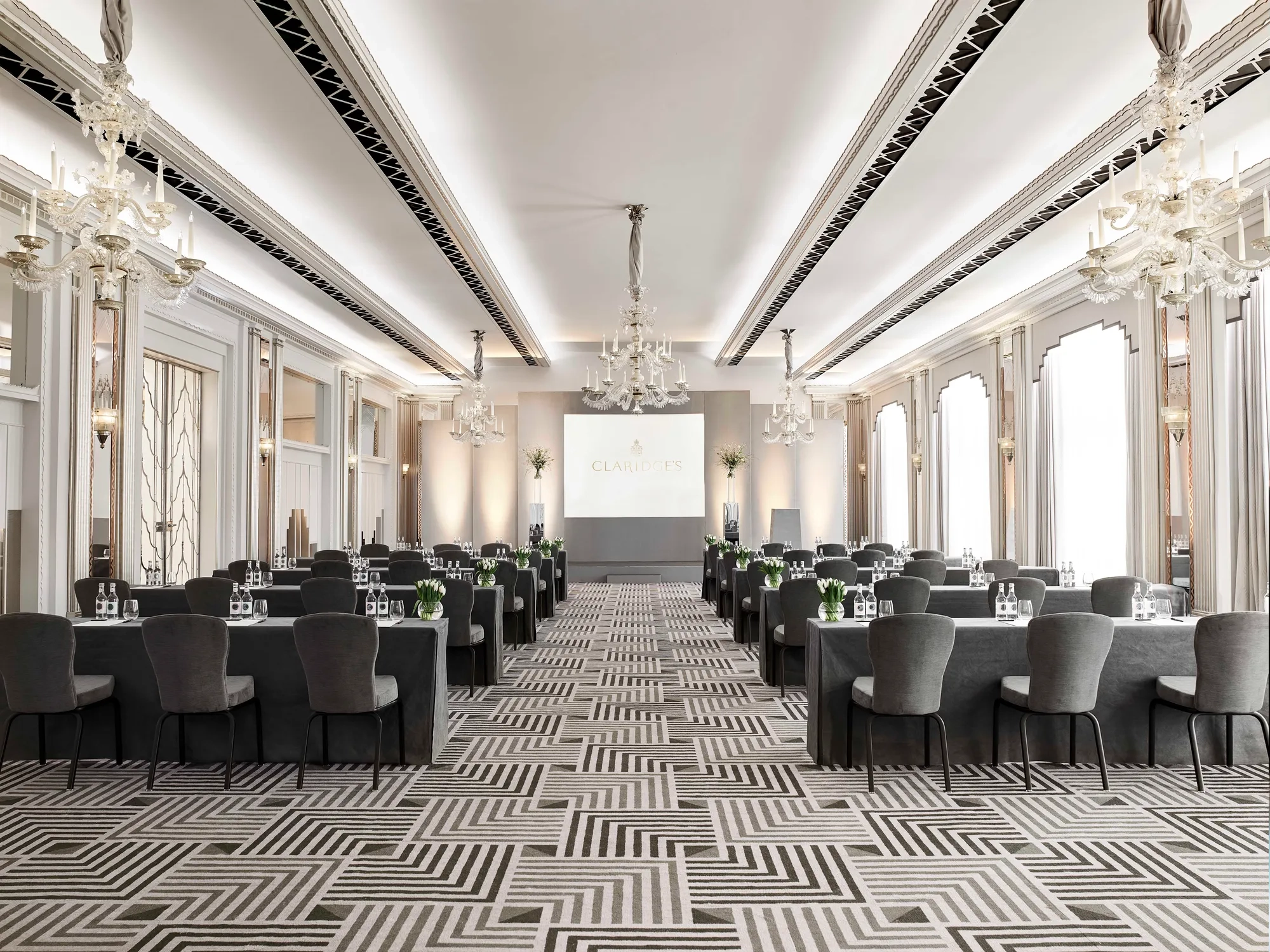 The Ballroom at Claridge’s arranged in a conference setup with long grey-covered tables and chairs facing a presentation screen, lit by chandeliers and large arched windows.