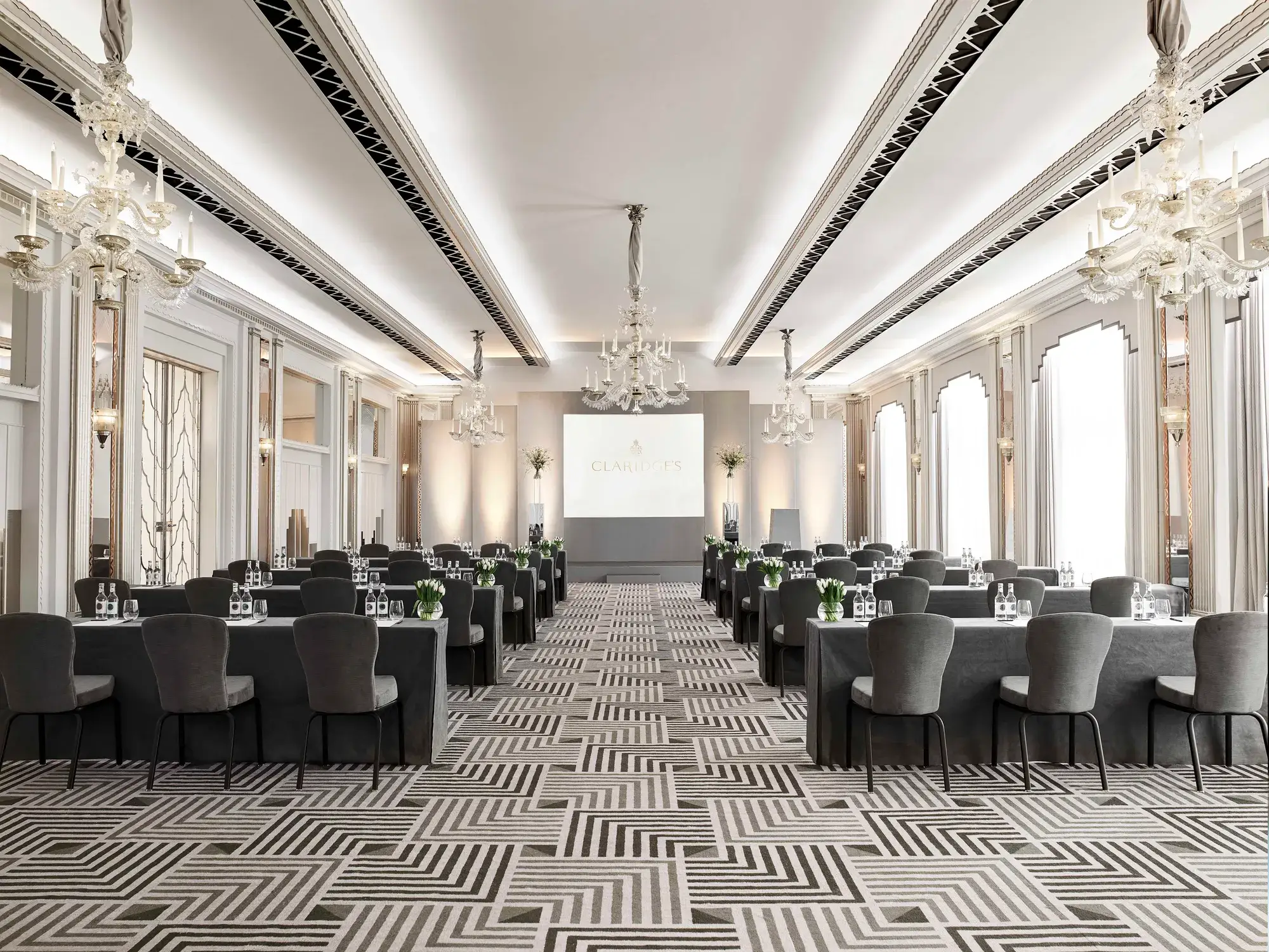 The Ballroom at Claridge’s arranged in a conference setup with long grey-covered tables and chairs facing a presentation screen, lit by chandeliers and large arched windows.