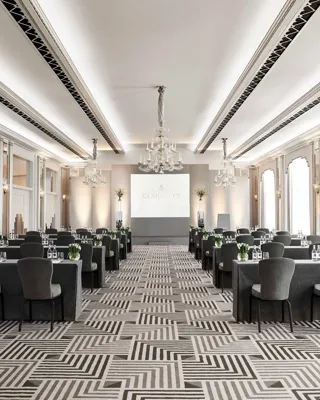 The Ballroom at Claridge’s arranged in a conference setup with long grey-covered tables and chairs facing a presentation screen, lit by chandeliers and large arched windows.