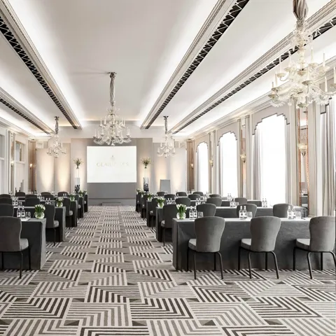 The Ballroom at Claridge’s arranged in a conference setup with long grey-covered tables and chairs facing a presentation screen, lit by chandeliers and large arched windows.