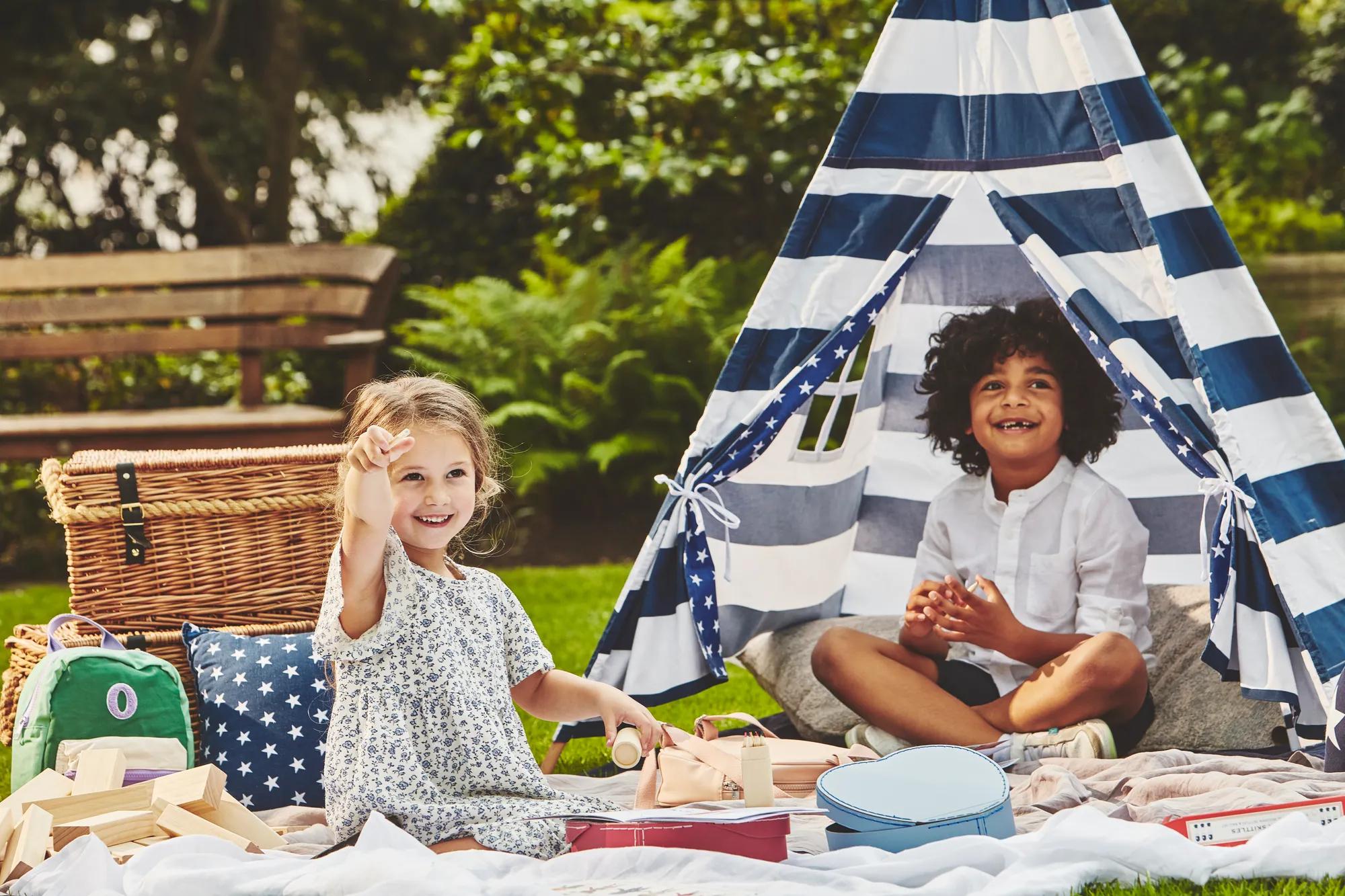 Two children play on a picnic blanket beside a striped teepee tent, surrounded by toys in a garden setting