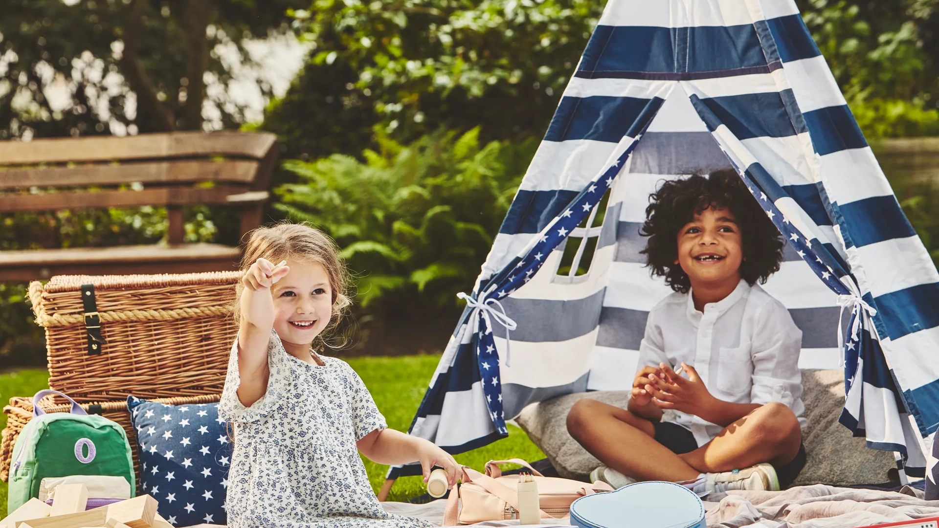 Two children play on a picnic blanket beside a striped teepee tent, surrounded by toys in a garden setting