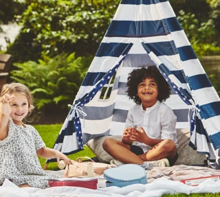 Two children play on a picnic blanket beside a striped teepee tent, surrounded by toys in a garden setting