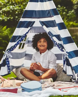 Two children play on a picnic blanket beside a striped teepee tent, surrounded by toys in a garden setting