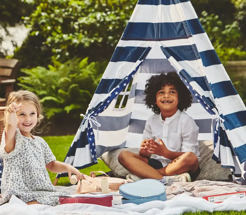 Two children play on a picnic blanket beside a striped teepee tent, surrounded by toys in a garden setting