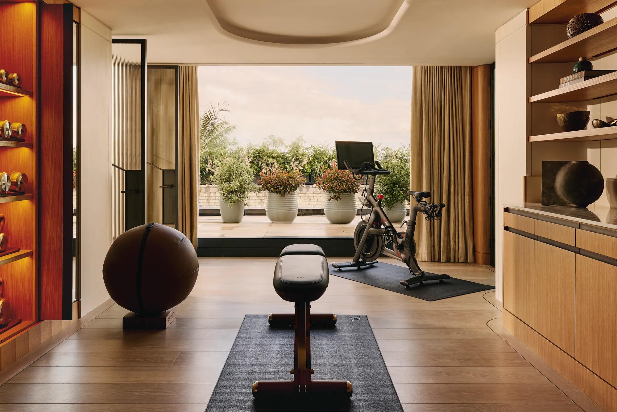A private in-suite gym with wood floors, free weights, a bench, and a Peloton bike, opening onto a terrace with potted greenery and a view of the sky.
