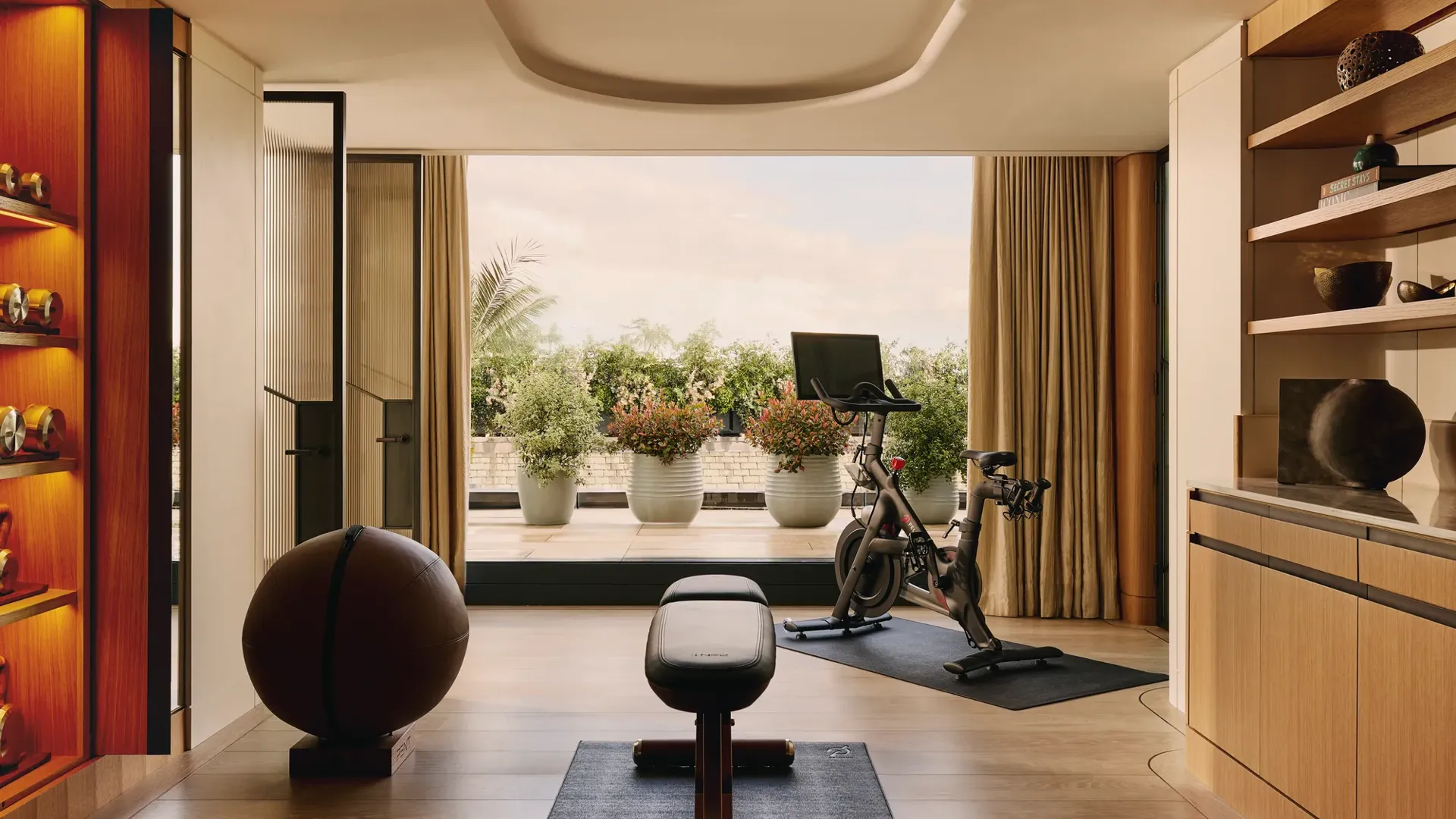 A private in-suite gym with wood floors, free weights, a bench, and a Peloton bike, opening onto a terrace with potted greenery and a view of the sky.