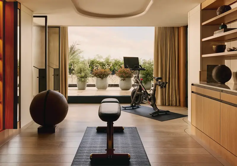 A private in-suite gym with wood floors, free weights, a bench, and a Peloton bike, opening onto a terrace with potted greenery and a view of the sky.