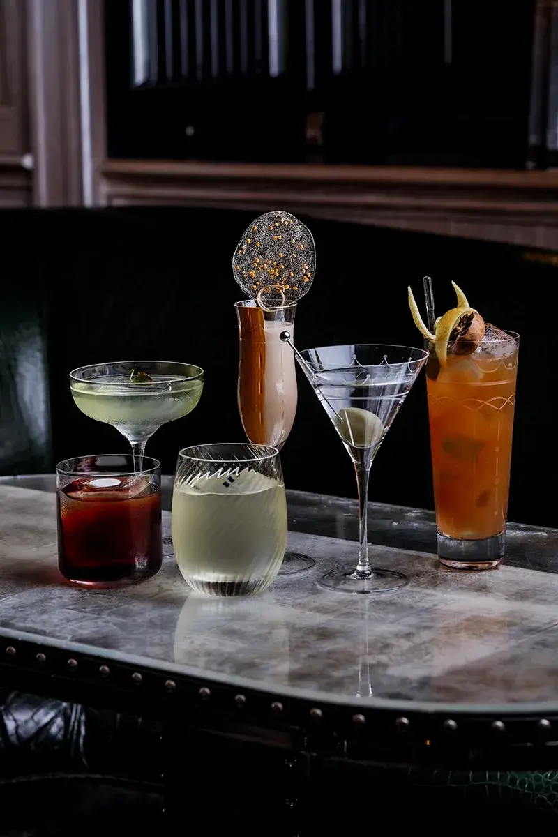 Six assorted cocktails on marble table in dimly lit bar, including martini, spritz, and coupe glasses