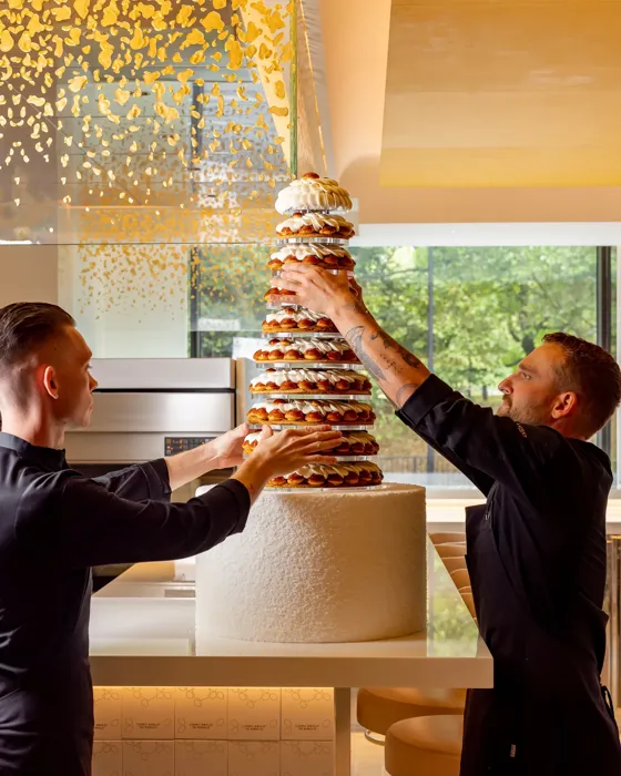 Pastry chefs assembling tall croquembouche wedding cake in modern kitchen with gold ceiling accents.