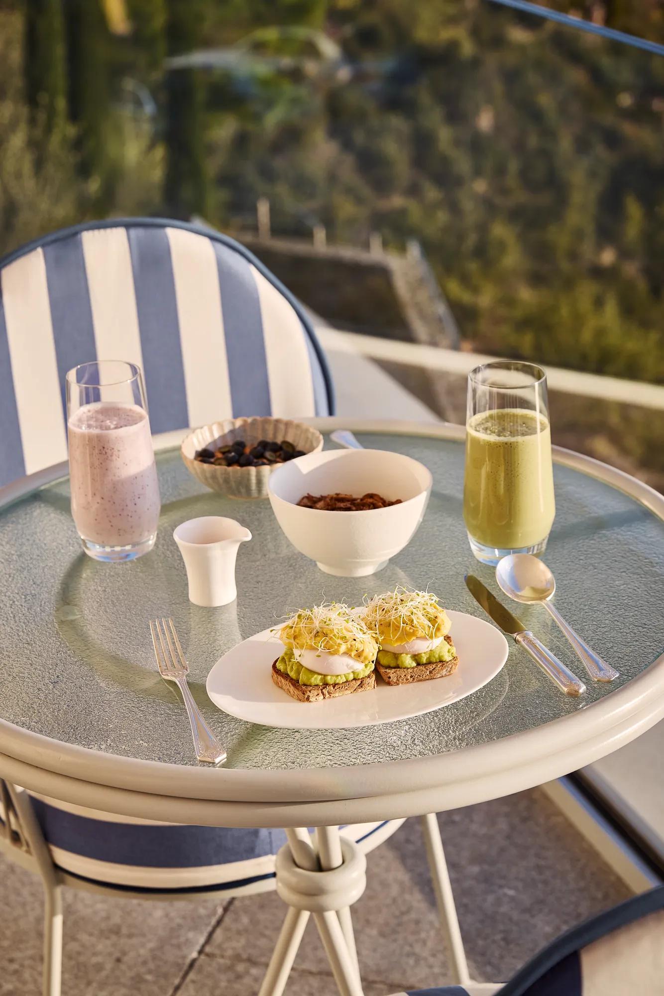 Breakfast on a terrace table with avocado toast, sprouts, bowls, smoothie, and green juice overlooking lush hillside views.