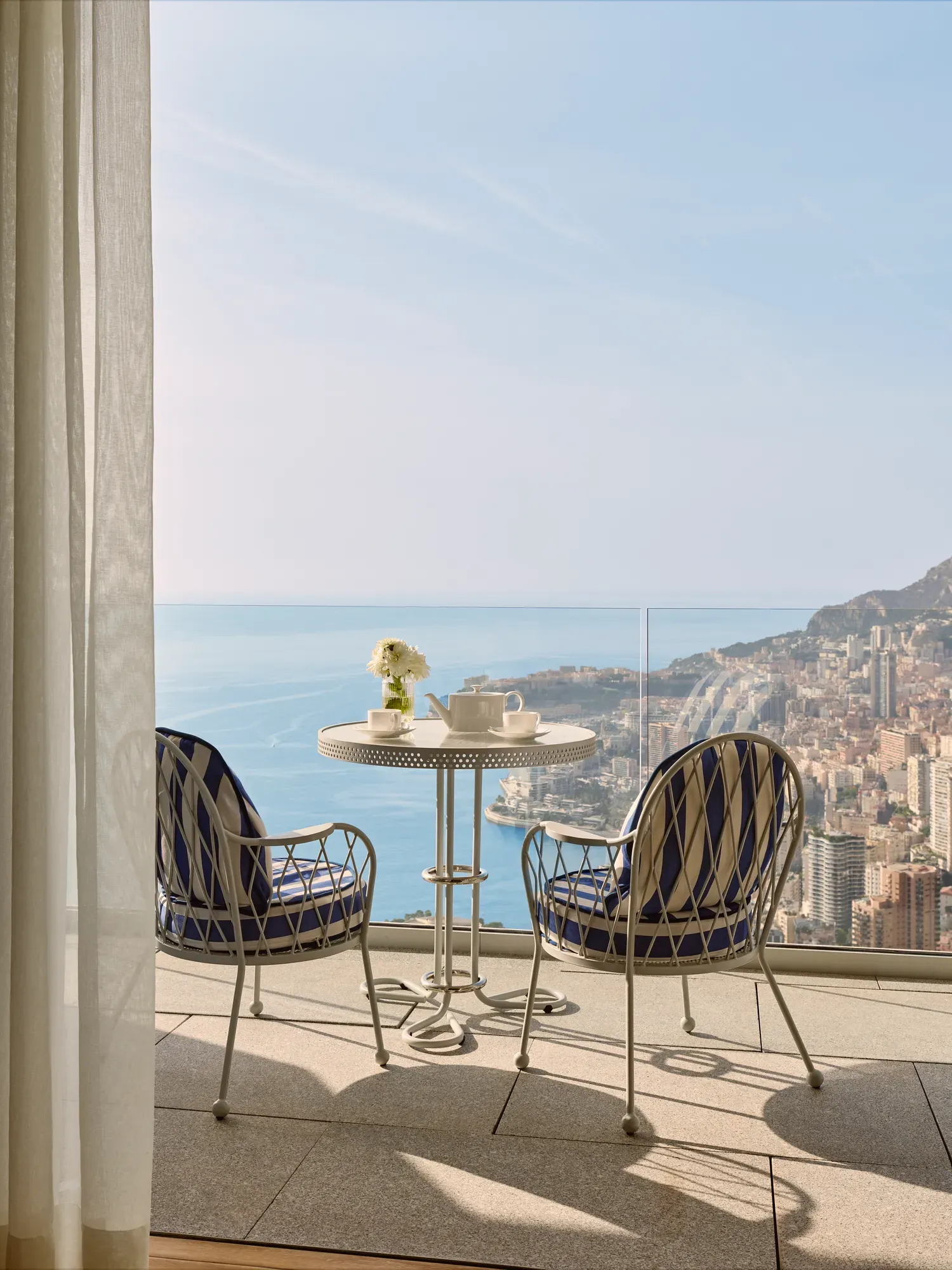 Balcony with a round table set for coffee and two blue-striped chairs, overlooking the city of Monte Carlo and the Mediterranean Sea.