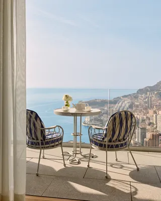 Balcony with a round table set for coffee and two blue-striped chairs, overlooking the city of Monte Carlo and the Mediterranean Sea.