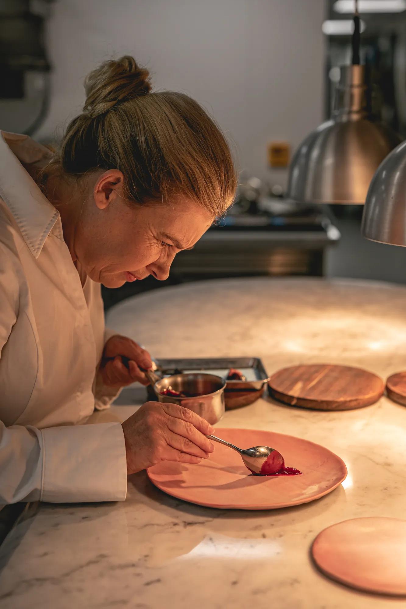 Chef plating dessert on pink plate, carefully spooning sauce with precision, lit by warm kitchen lighting and surrounded by utensils.
