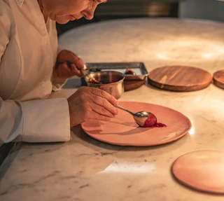 Chef plating dessert on pink plate, carefully spooning sauce with precision, lit by warm kitchen lighting and surrounded by utensils.