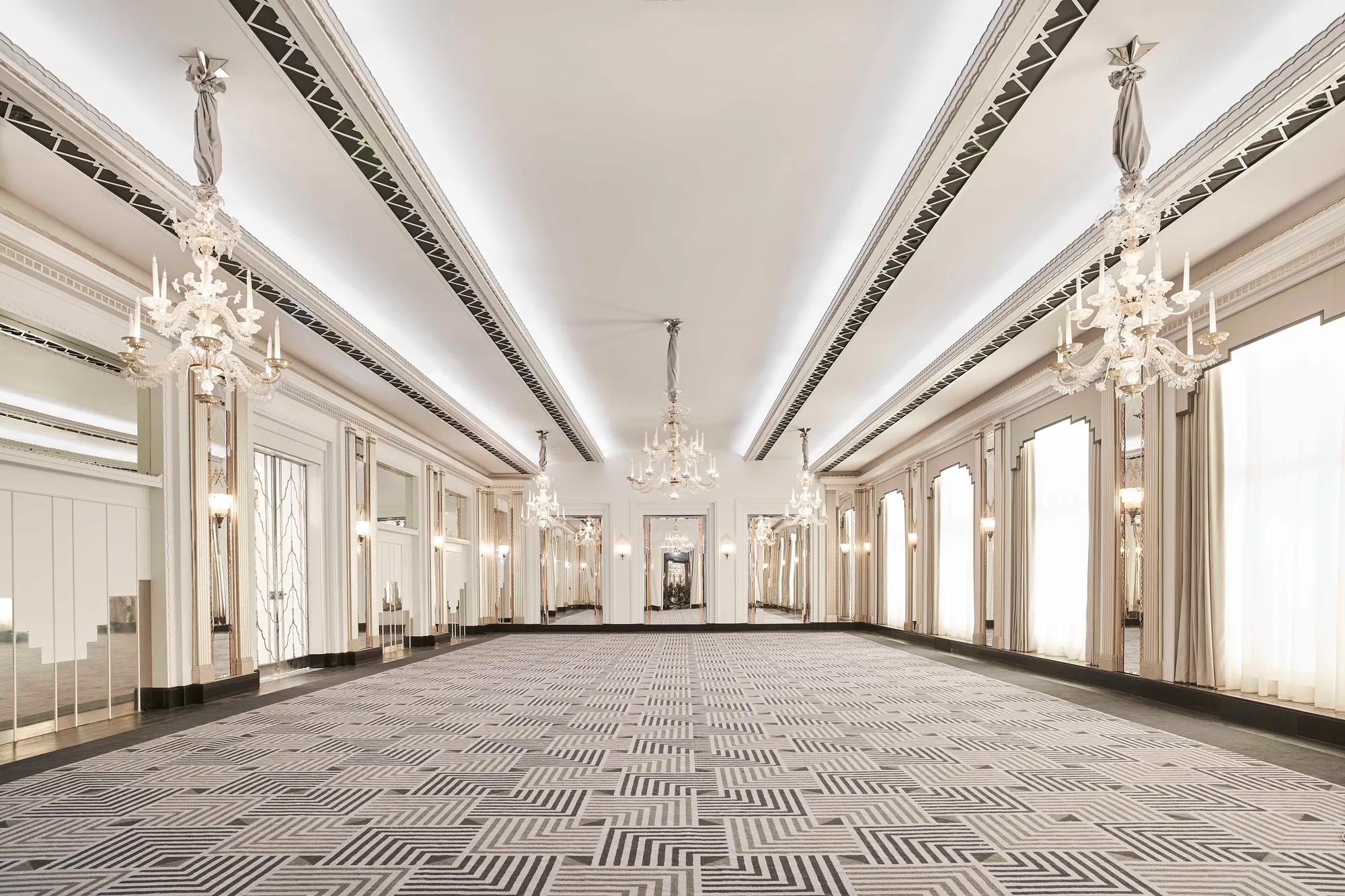 The Ballroom at Claridge’s shown empty, featuring Art Deco chandeliers, mirrored walls, tall arched windows with draped curtains, and a geometric patterned carpet in soft grey tones.