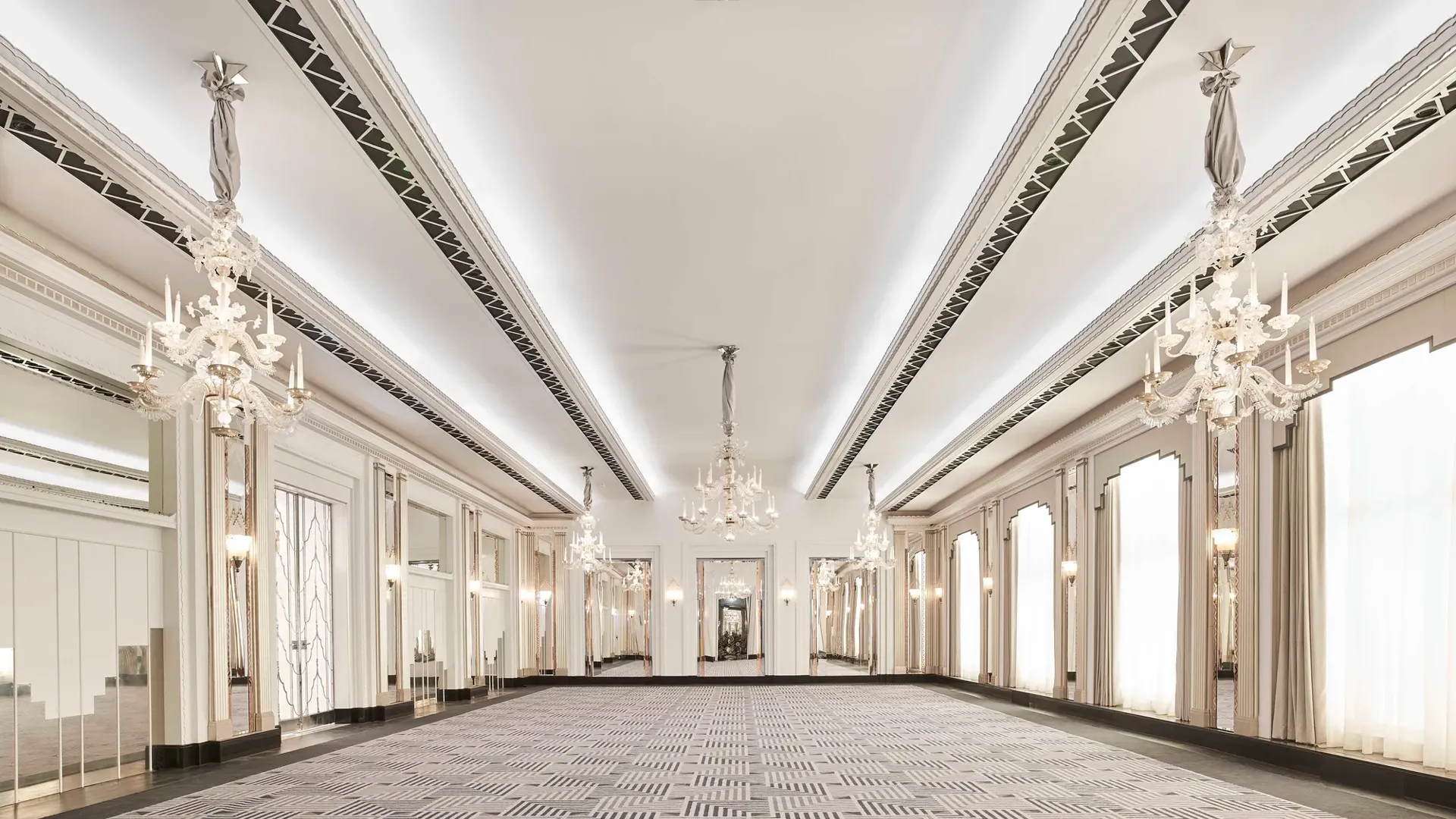 The Ballroom at Claridge’s shown empty, featuring Art Deco chandeliers, mirrored walls, tall arched windows with draped curtains, and a geometric patterned carpet in soft grey tones.