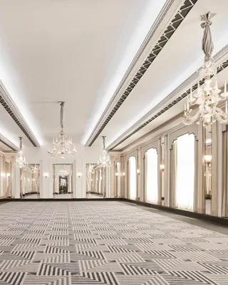 The Ballroom at Claridge’s shown empty, featuring Art Deco chandeliers, mirrored walls, tall arched windows with draped curtains, and a geometric patterned carpet in soft grey tones.
