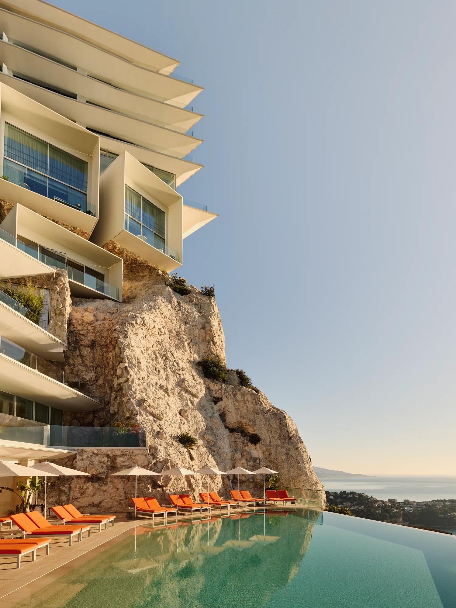 Clifftop hotel with an infinity pool and orange loungers overlooking the sea under a clear sky.