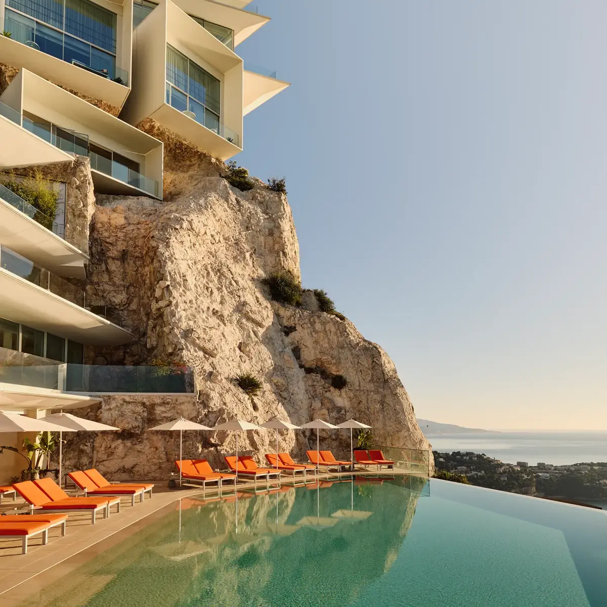 Hôtel perché sur une falaise avec piscine à débordement et transats orange surplombant la mer sous un ciel clair.