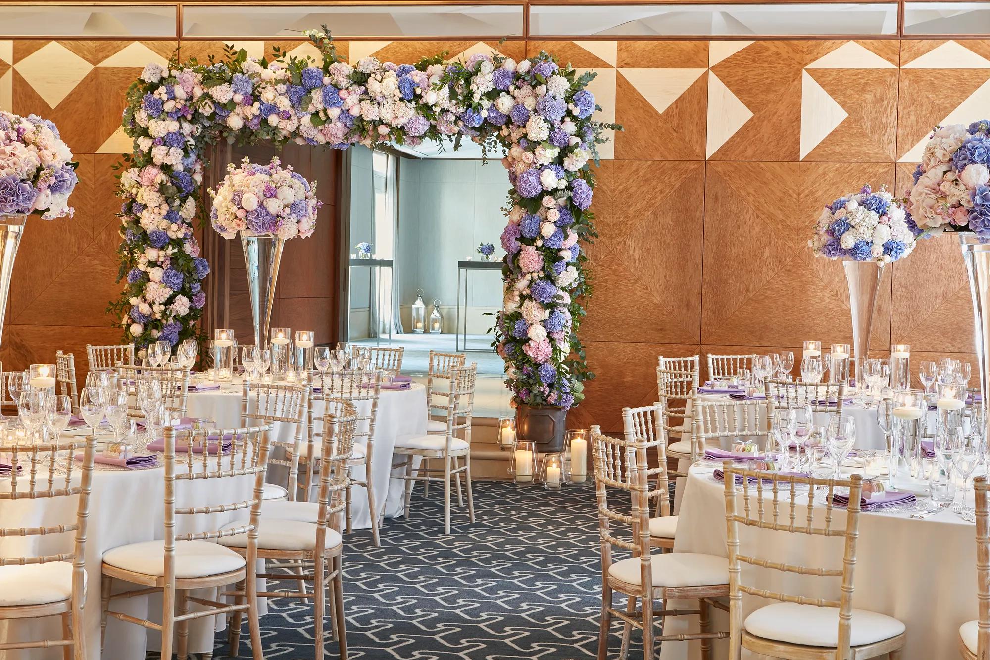 Large, elegant ballroom with round tables dressed in white linens and tall gold chairs, centred on a dramatic floral arch of purple, blue, and white hydrangeas and roses, surrounded by candlelit glass votives