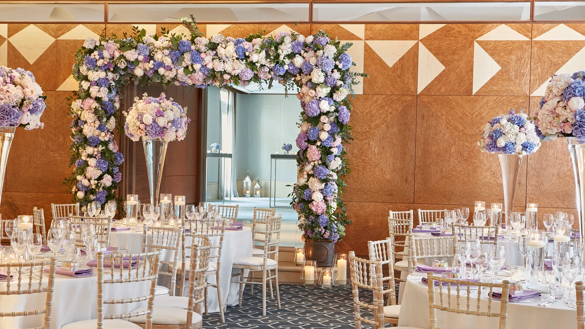 Large, elegant ballroom with round tables dressed in white linens and tall gold chairs, centred on a dramatic floral arch of purple, blue, and white hydrangeas and roses, surrounded by candlelit glass votives